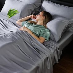 a woman laying on top of a bed covered in gray sheets and pillows next to a green plant