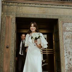 a woman in a white dress is walking out of a building with a man wearing a suit and tie