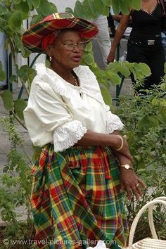 an older woman wearing a plaid skirt and hat standing in front of some plants with her hands on her hips