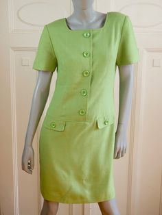 This 1990s vintage green dress has a round collarless neckline and features five decorative buttons on the front from the neckline to the dropped waistline. There are also two decorative buttons on each of the two faux pockets on the front. The dress has padded shoulders, an above-the-knee hemline, and a long back zipper that makes dressing an ease. It is fully lined in a green satin polyester fabric. Bust = 38 inches (96cm) Shoulder Width = 16 inches (40.64cm) Waist = 32 inches (81.28cm) Hips = Retro Fitted Dresses With Buttons, Classic Green Spring Dress, Fitted Knee-length Vintage Dress With Buttons, Classic Green Knee-length Dress, Vintage Green Dress For Work, Classic Green Dress With Buttons, Retro Green Dress For Work, Fitted Green Vintage Dress, Classic Vintage Dress With Buttons For Spring