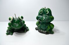 two green frog figurines sitting next to each other on a white table top