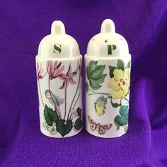 two ceramic salt and pepper shakers with flowers painted on the sides, sitting on a purple cloth