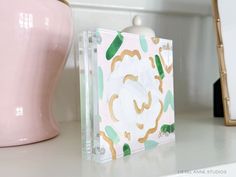 a pink vase sitting on top of a white counter next to a clear box with gold foil