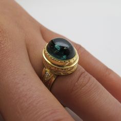 This exquisite, regal-looking dome ring features a fine, 9.80 carat oval green tourmaline cabochon set in a beautifully handmade, hand engraved bezel. The tourmaline has a rich, bluish green body color and has excellent clarity and transparency. The ring was designed to showcase this gorgeous gem, with a brilliant white diamond on either side of the center stone for a touch of added luxury. The handsome bezel has a multi-layered appearance and is as beautiful from the side as from the top. Intri Luxury Emerald Ring With Oval Cabochon, Green Gemstone Cabochons For Formal Use, Formal Fine Jewelry Domed Cabochons, Formal Domed Cabochons Fine Jewelry, Luxury Green Cabochons, Luxury Oval Gemstone Cabochons, Luxury Round Cabochons With Bezel Setting, Elegant Domed Emerald Ring Gift, Luxury Cabochons With Bezel Setting