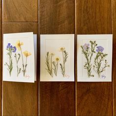 three cards with pressed flowers on them sitting on a wooden table next to each other