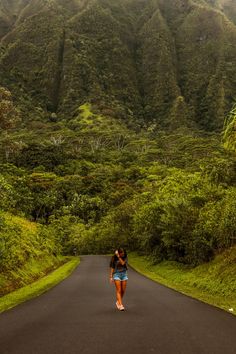 a woman walking down the middle of a road in front of green mountains and trees