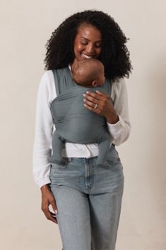 a woman is holding a baby in a grey sling and smiling at the camera while wearing jeans