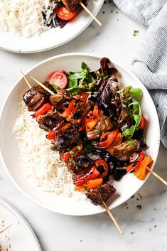 two plates filled with meat and veggies on skewers next to rice