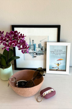 purple flowers in a pink bowl next to a pair of sunglasses and a framed photograph