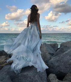 a woman is standing on some rocks by the ocean with her dress blowing in the wind