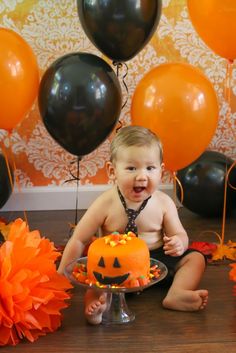 a baby is sitting on the floor in front of a cake and balloons that look like pumpkins