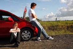 two people sitting on the side of a road next to a red car with its door open