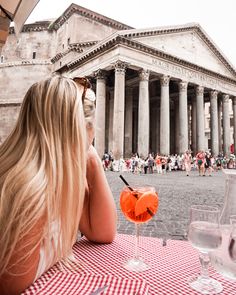 a woman sitting at a table with an orange drink in front of a large building