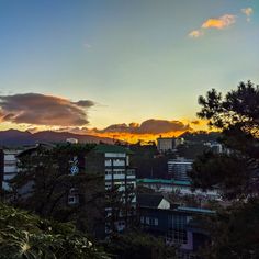 the sun is setting over some buildings and trees with mountains in the backround