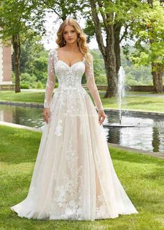 a woman wearing a wedding dress with long sleeves and flowers on the skirt is standing in front of a fountain