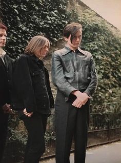 three people standing next to each other in front of a wall with ivy growing on it