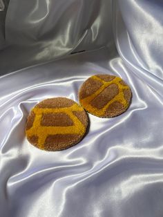 two brown and yellow coasters sitting on top of a white satin covered bed sheet