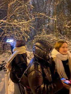 three people standing in the snow looking at their cell phones, one woman is wearing a scarf