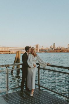 a man and woman standing next to each other on a pier near the water with a bridge in the background