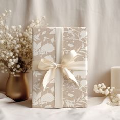 a gift box wrapped in white paper with a bow on it next to some flowers