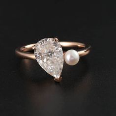 an engagement ring with a pear shaped diamond and a pearl on the side, sitting on a black surface