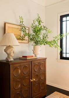 a wooden cabinet with two vases on top and a lamp next to it in front of a window