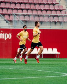 two soccer players run on the field during a training session in front of an empty bleachers