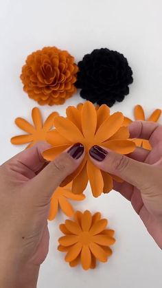 two hands are holding orange and black paper flowers on a white surface, with one hand reaching for the flower