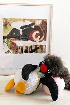 a stuffed penguin sitting next to an art piece on a table with a framed photo in the background