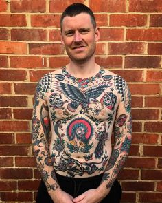 a tattooed man standing in front of a brick wall with his hands on his hips