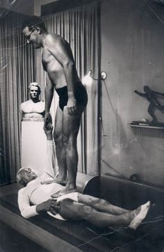 an old black and white photo of a man standing over another man laying on the bed