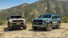 two toyota trucks parked in the dirt near some mountains and trees, one is gray