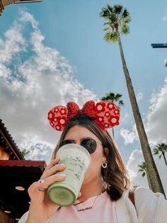 a woman drinking from a cup with minnie ears on her head and wearing sunglasses in front of palm trees