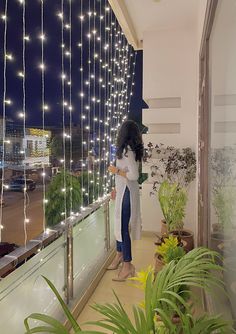 a woman standing on a balcony next to a window covered in fairy lights and potted plants