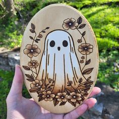 a hand holding up a carved wooden plaque with a ghost on it's face