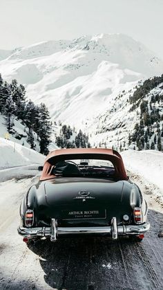 an old black car parked on the side of a road in front of snow covered mountains