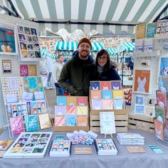 The photograph shows a man and a woman standing in thick winter layers and smiling, behind a market table stall with temporary walls attached at either side, to a green & white stripy gazebo. The products on display are brightly coloured cards, calendars, notebooks, prints, mugs and tote bags, with illustrations of dogs, cats, rabbits, goldfish, hamsters and more! Craft Fair Display Table, Booth Display Ideas Diy, Craft Fair Table, Christmas Market Stall, Market Display