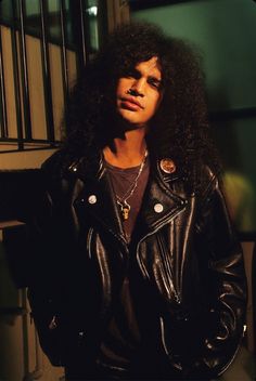 a man with long hair wearing a black leather jacket and standing in front of a building