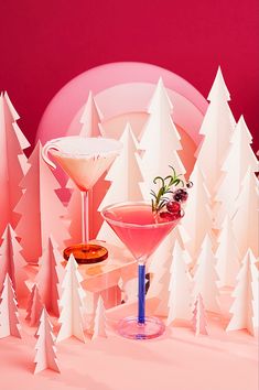 two martini glasses with garnish sit on a pink surface surrounded by white trees