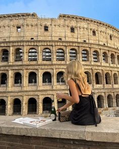 a woman sitting on the ledge in front of an arena looking at her cell phone