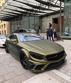 a gold colored mercedes benz coupe parked in front of a building with people walking by