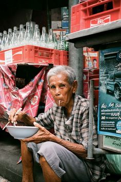 an old man sitting on the ground eating from a bowl with chopsticks in his mouth