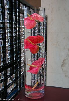 three pink flowers in a clear vase on a table next to many stacks of files