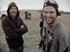 two men standing next to each other in a field with motorcycles behind them and one man wearing a hoodie