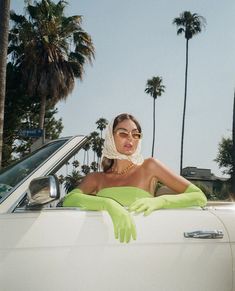 a woman wearing green gloves sitting in the back of a white car with palm trees behind her