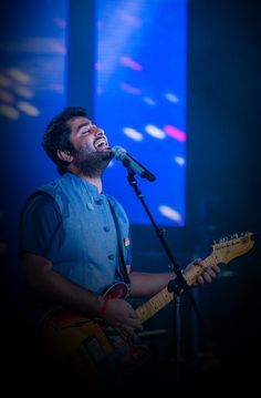 a man singing into a microphone while holding a guitar in front of him on stage