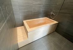 a wooden bathtub in a bathroom with grey tile walls and flooring, along with a shower head