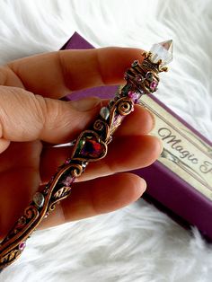 a hand holding an ornate gold and pink colored piece of jewelry in front of a purple box