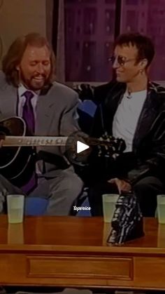 two men are sitting on a couch playing guitars and drinking beverages in front of them