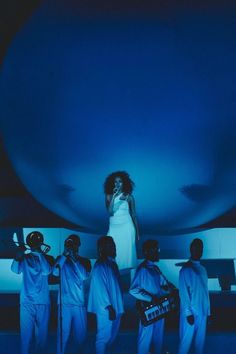 a woman standing on top of a stage surrounded by other people in white outfits and blue lighting